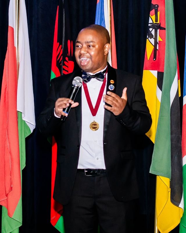 Frank Tsuro in black tie, speaking into a handheld microphone in front of a row of national flags. Colour.
