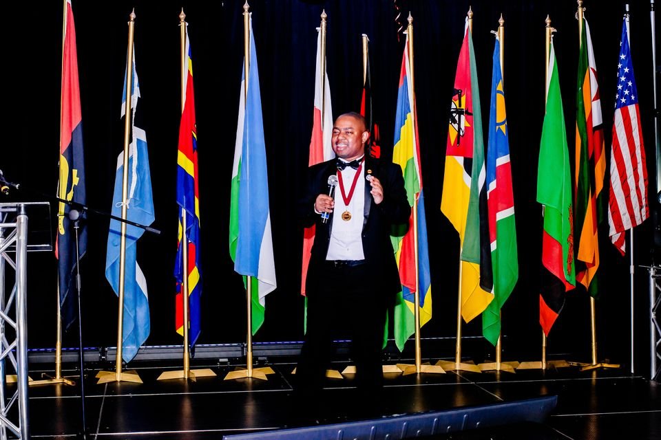 Frank Tsuro in black tie on a stage lined with national flags. Wide frame in colour.