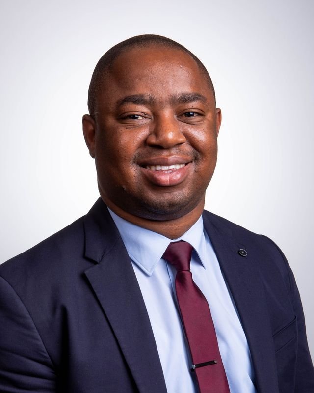 Frank Tsuro in a navy suit, light blue shirt and maroon tie, turned slightly to the right. Studio headshot in colour.
