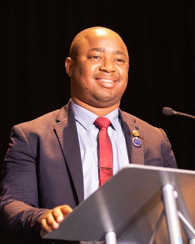 Frank Tsuro smiling at a lectern in a navy suit and red tie. Colour.