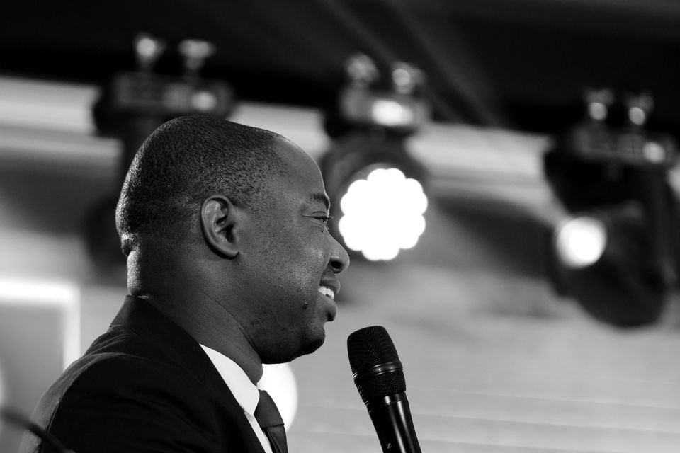 Frank Tsuro in profile at a microphone under a row of stage lights. Wide frame in black and white.