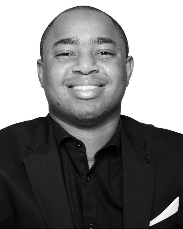 Frank Tsuro laughing in a dark jacket, black shirt and white pocket square. Studio portrait in black and white.